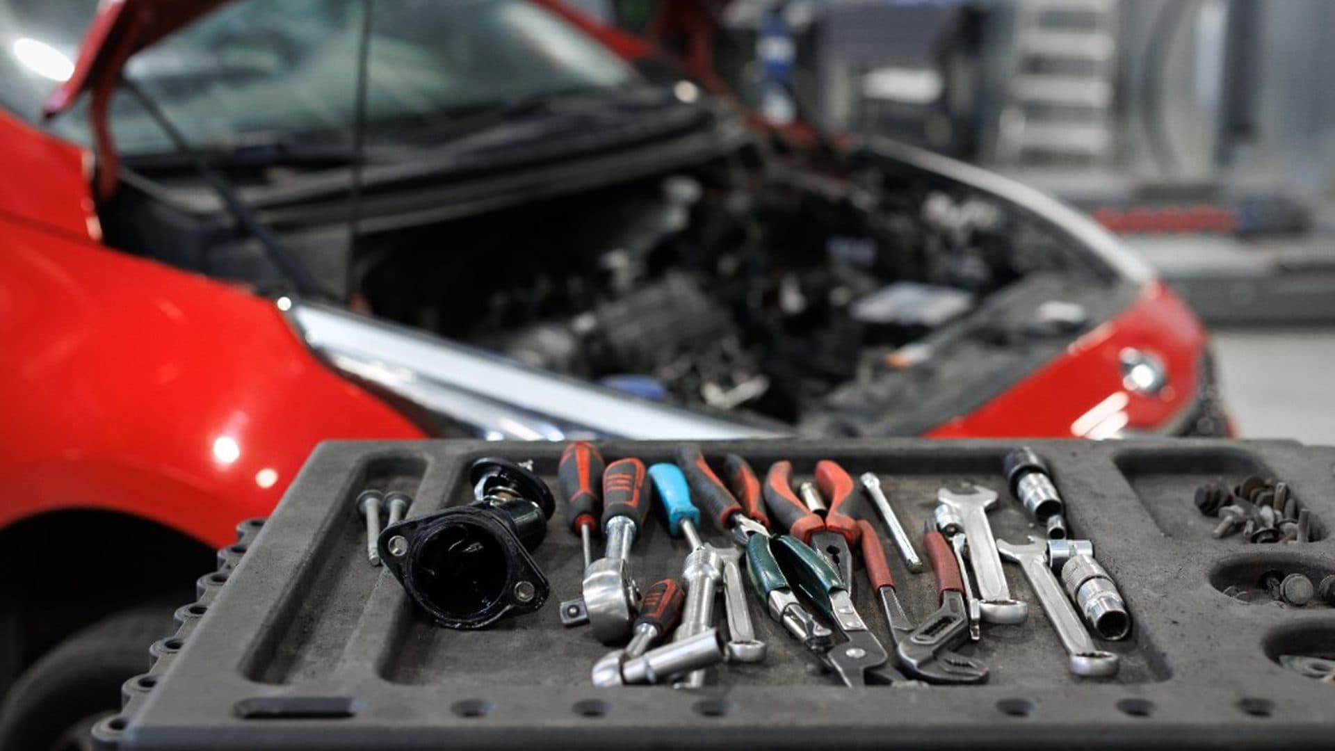 Hand tools on a service tray with a red car hood open in the garage behind them